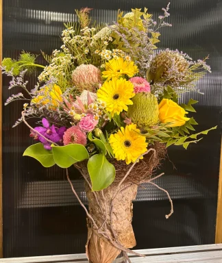 "Rustic Heaven floral arrangement: Gerberas, orchids, Protea, roses, banksia leaves, and chrysanthemums in a unique vase wrapped in lotus leaves, with an earthy charm and subtle mystic accents."