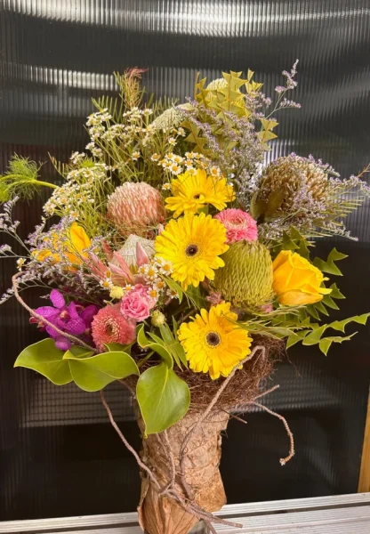 "Capture the essence of nature with 'Rustic Heaven' floral arrangement. Dried lotus leaf-wrapped vase holds fresh Gerberas, orchids, Protea, yellow roses, banksia leaves, and playful pom pom chrysanthemums. A rustic design with mystic and chamomile accents for an earthy, captivating display."